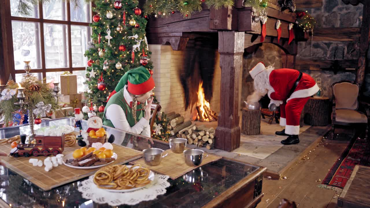 Santa Claus and an elve girl. Santa preparing tea. Santa Claus with boiling teapot, Curious girl wears elves clothes. Happy elf girl in fairy costume sitting near the table. Santa near Christmas fireplace.