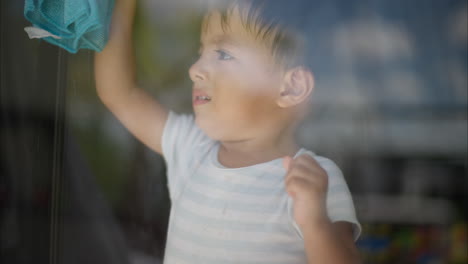 Lindo-Bebé-Latino-Limpiando-La-Ventana-Haciendo-Sus-Tareas-Usando-Un-Paño-Azul-En-Una-Mañana-Cálida-Y-Soleada