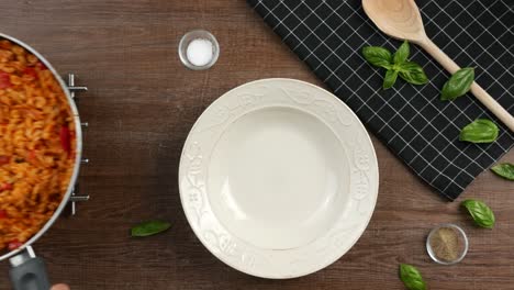 top down shot of a hot pan with creamy fusilli pasta with tomato sauce appearing on wire rack, situated on wooden table decorated with white deep plate, wooden spatula, black cloth and basil leaves