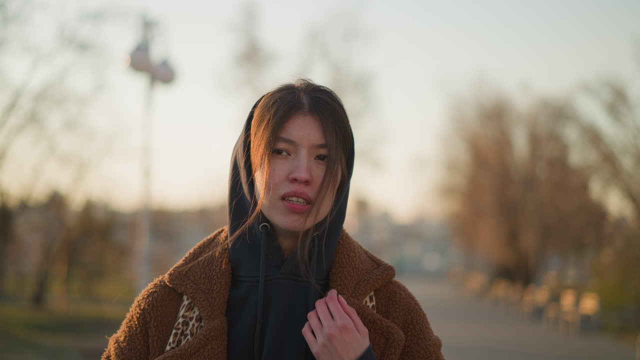 황혼한 소녀가 해가 지는 동안 외로운 공원을 고, 갈색 코트와 후디를 입고, 머리카락이 부분적으로 얼굴을 고 있습니다.