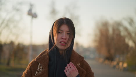 a downhearted girl walking through a lonely park at sunset, wearing a brown coat and a hoodie with her hair partially covering her face. somber mood as she appears deep in thought