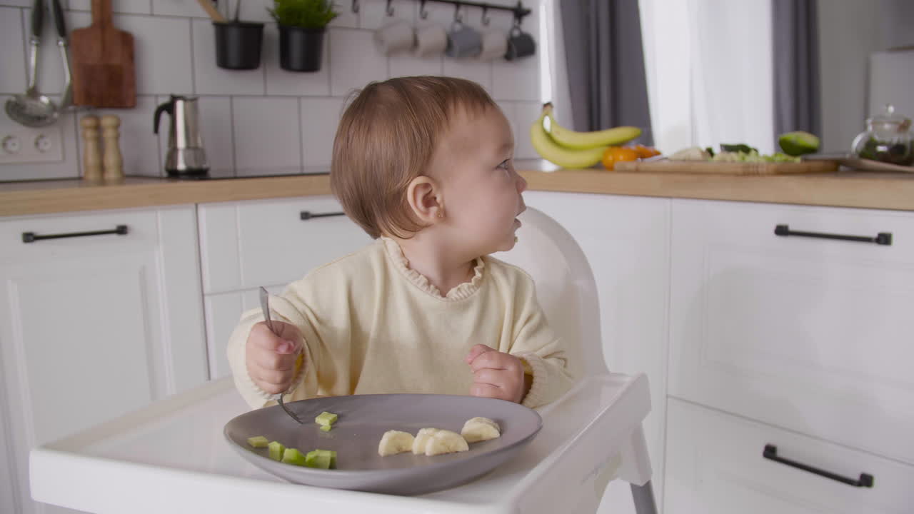 Cute Baby Girl Holding Fork And Looking Around While Sitting In Her High Chair In The Kitchen And Eating Fruit