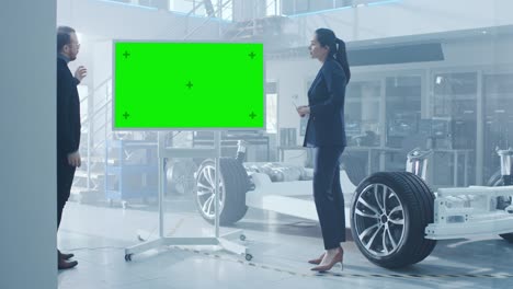 male and female design engineers are discussing something on a interactive whiteboard with green screen next to an electric car chassis prototype. in high tech laboratory facility with vehicle frame.