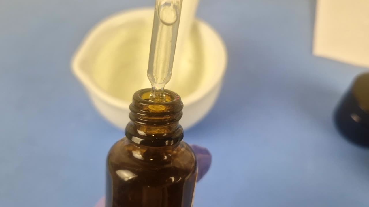 Macro shot of a clear cosmetic serum dripping from a glass dropper into an amber bottle. Shows pharmacist's gloved hands in a clean laboratory, highlighting precision