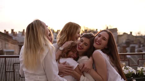 Attractive-caucasian-girls-are-hugging-standing-outside-on-a-terrace-or-balcony.-Six-beautiful-young-women-in-white-shirts-and-with-red-lipstick.-Hen-party.-Evening-dusk.-Love-and-friendship
