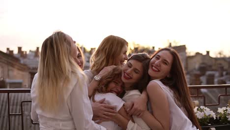 attractive caucasian girls are hugging standing outside on a terrace or balcony. six beautiful young women in white shirts and with red lipstick. hen party. evening dusk. love and friendship