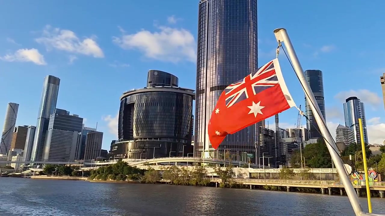 Vlajka Austrálie vlající na lodi s panoramatem města Brisbane v pozadí. Malebný výhled na řeku s moderními budovami pod jasnou modrou oblohou, zachycující cestování a národní identitu v Austrálii.