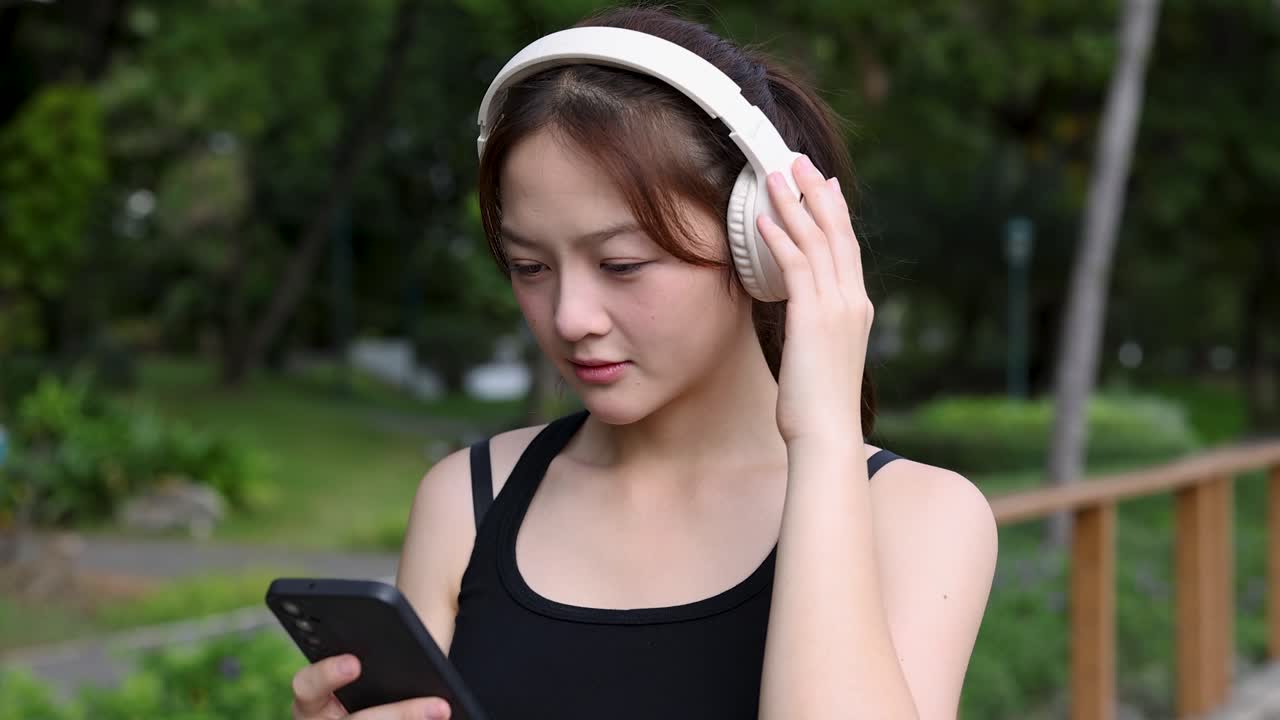 Una giovane donna in abbigliamento sportivo usa uno smartphone mentre indossa le cuffie in un parco.