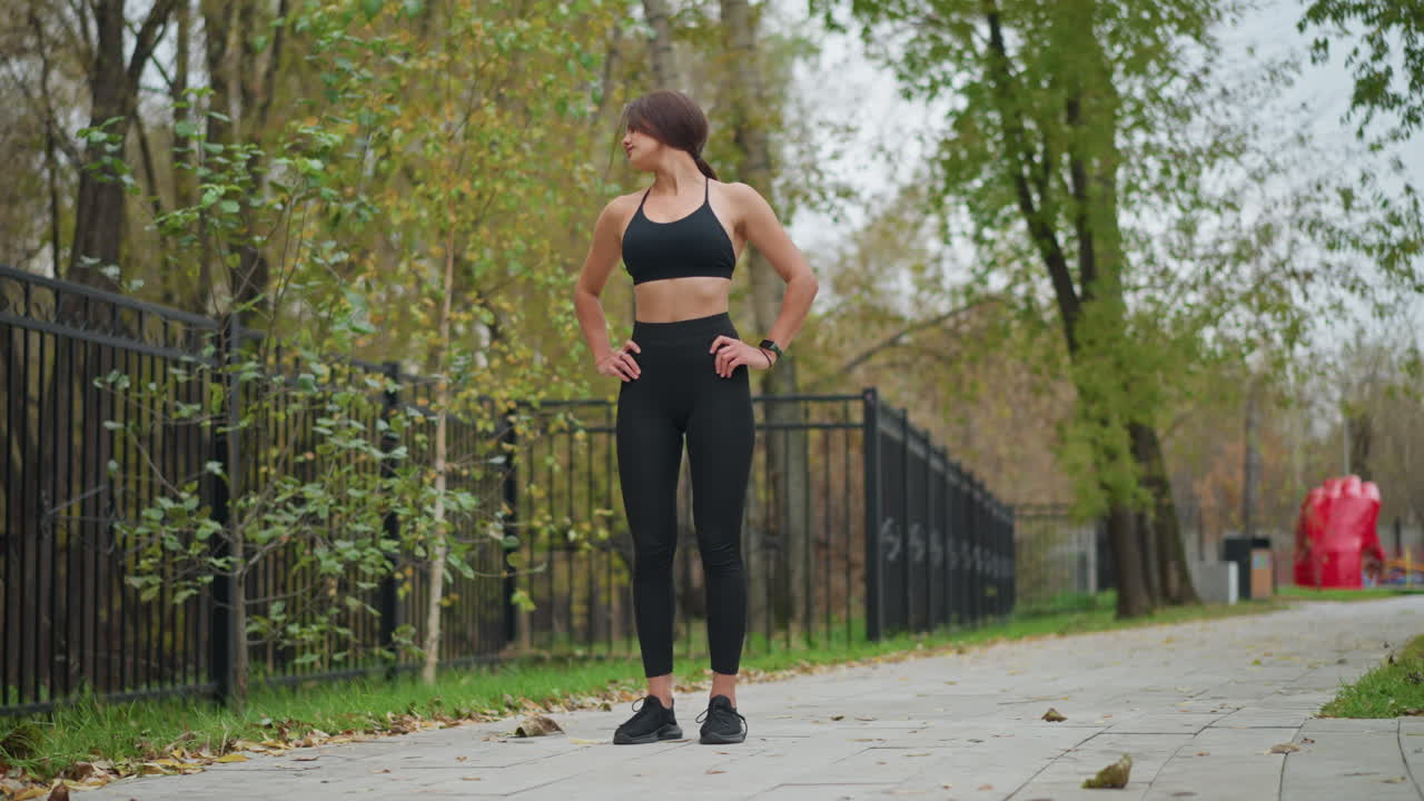 Lady in black neck top and leggings standing, stretching neck with hand on waist in park, blurred background featuring iron fence and trees, outdoor fitness, neck flexibility, posture correction
