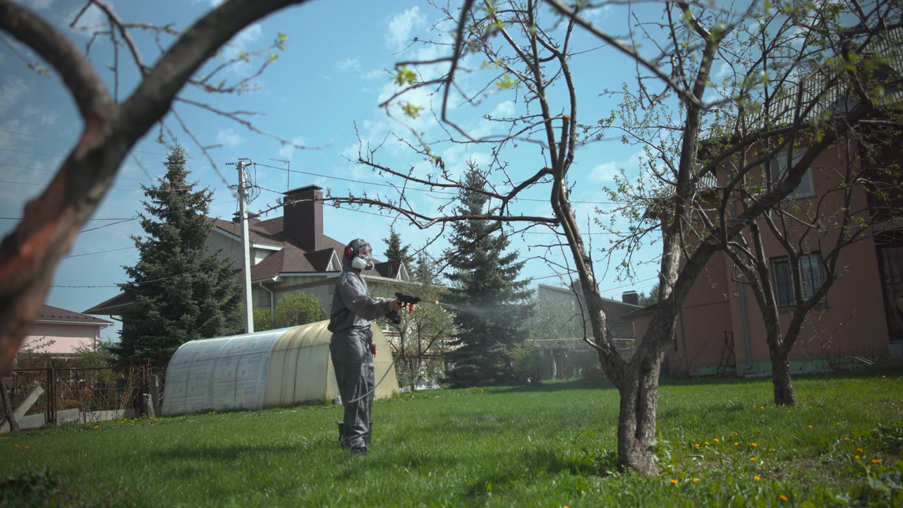 A person wearing a reflective protective suit stands on a green lawn in a sunny residential yard, with leafing trees, a fence, and houses visible in the