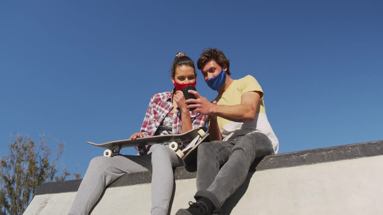 Donna e amico caucasici che indossano mascherine, seduti e che usano lo smartphone.