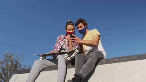 Caucasian-woman-and-man-friend-wearing-face-mask,-sitting-and-using-smartphone