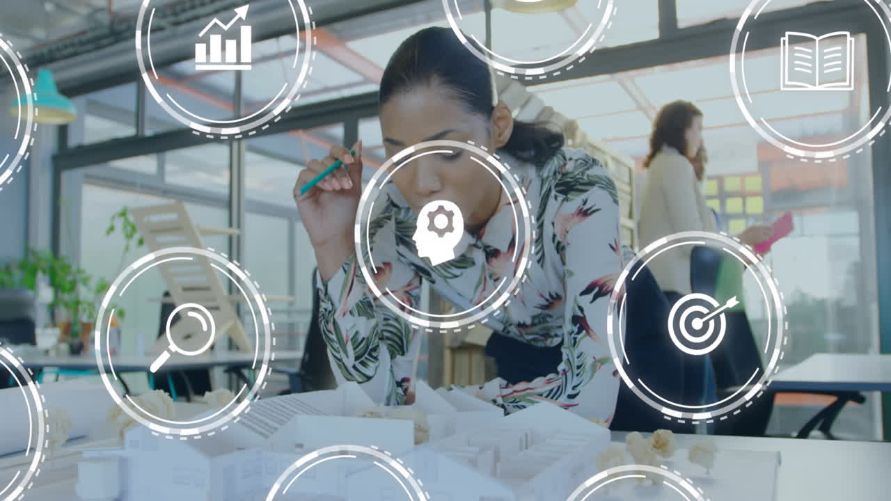 animación de múltiples iconos digitales contra una mujer biracial trabajando en un plan modelo en la oficina