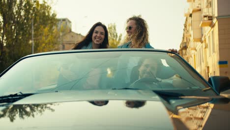 Vorderansicht:-Blondes-Mädchen-Mit-Schwarzer-Brille-In-Einem-Grauen-Cabrio-Tanzt-Und-Hat-Spaß-Neben-Ihren-Freunden