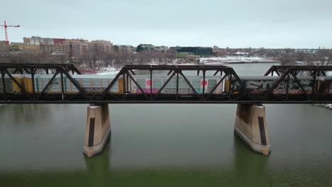 Close-Up-4K-Winter-Snow-Covered-Freezing-Northern-Landscape-Drone-Shot-Iron-Steel-Train-Industrial-Vehicle-Bridge-Over-the-Assiniboine-Red-River-Green-Water-in-Winnipeg-Manitoba-Canada