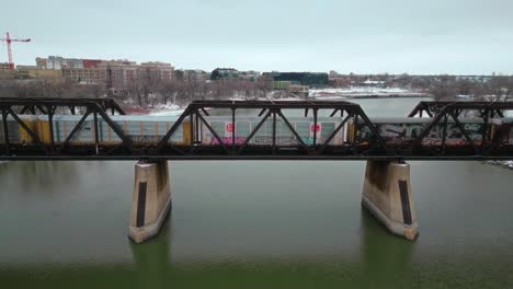 close up 4k winter snow covered freezing northern landscape drone shot iron steel train industrial vehicle bridge over the assiniboine red river green water in winnipeg manitoba canada