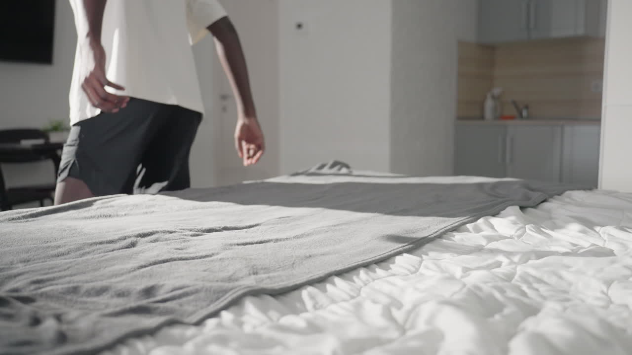 Person in white shirt smoothing out grey blanket on bed, adjusting fabric to ensure smoothness, soft natural light brightening modern bedroom, minimalist interior, peaceful and organized environment