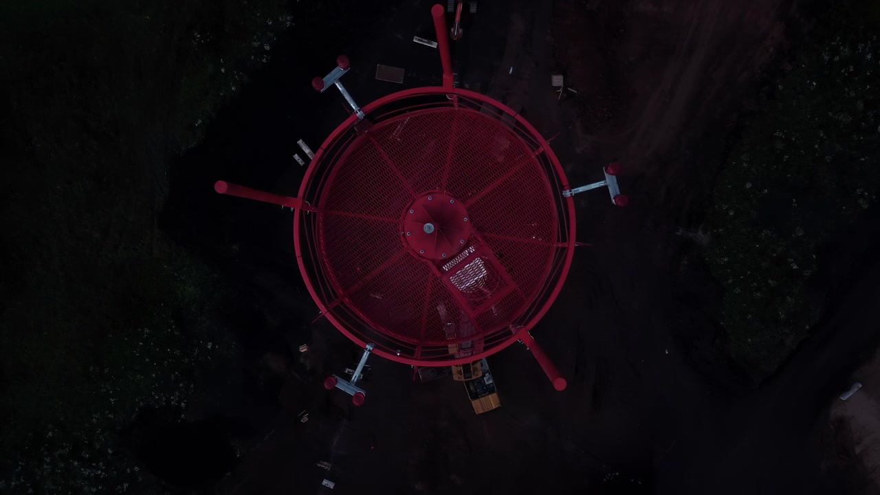 Aerial overhead view of a centered red circular industrial structure with metal supports, geometric rings, and strong contrast against a dark background under