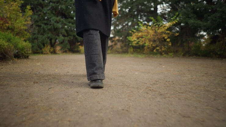 Close-up back view of feet in black jeans and canvas shoes walking along an autumn garden path, surrounded by colorful foliage and peaceful nature