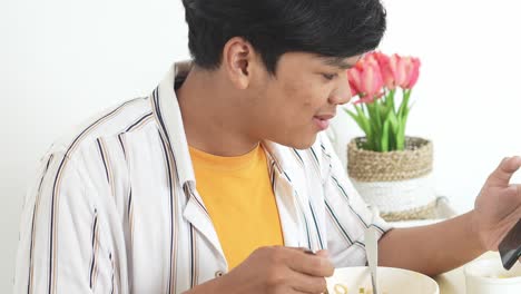 Joven-Asiático-Comiendo-Fideos-Instantáneos-Y-Charlando-Por-Teléfono-Inteligente-Con-Un-Amigo-En-Casa