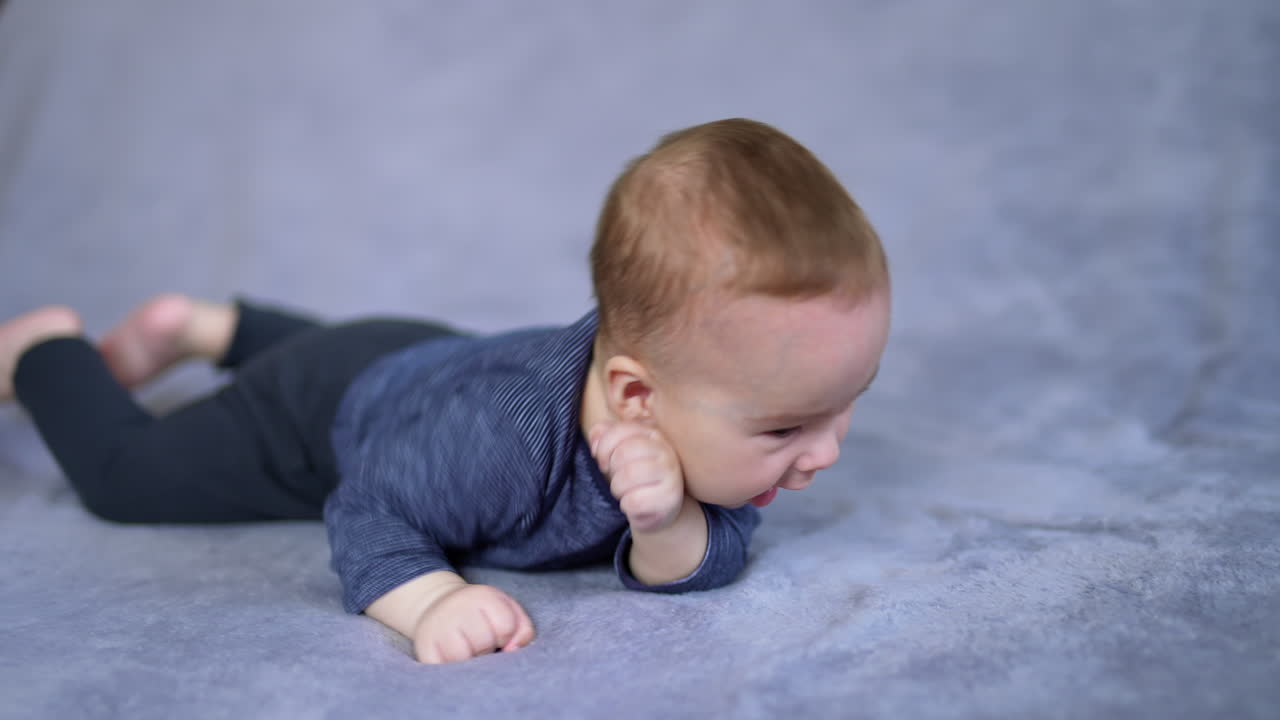 Active little baby boy turning from his back onto belly. Healthy child in blue clothes lies on the grey background. Close up.