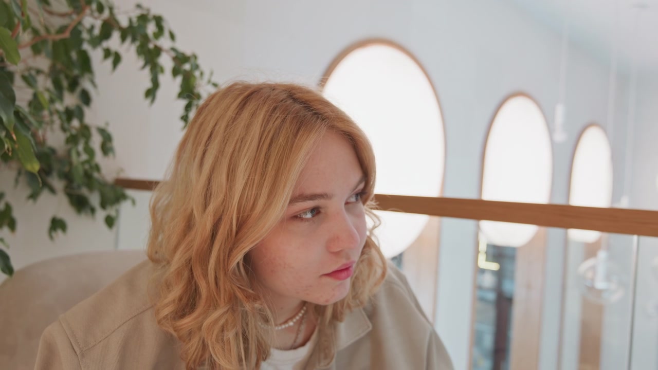 Close up of young woman with wavy blonde hair wearing beige jacket and white top seated in indoor setting. scene features soft lighting from large round windows behind her green foliage