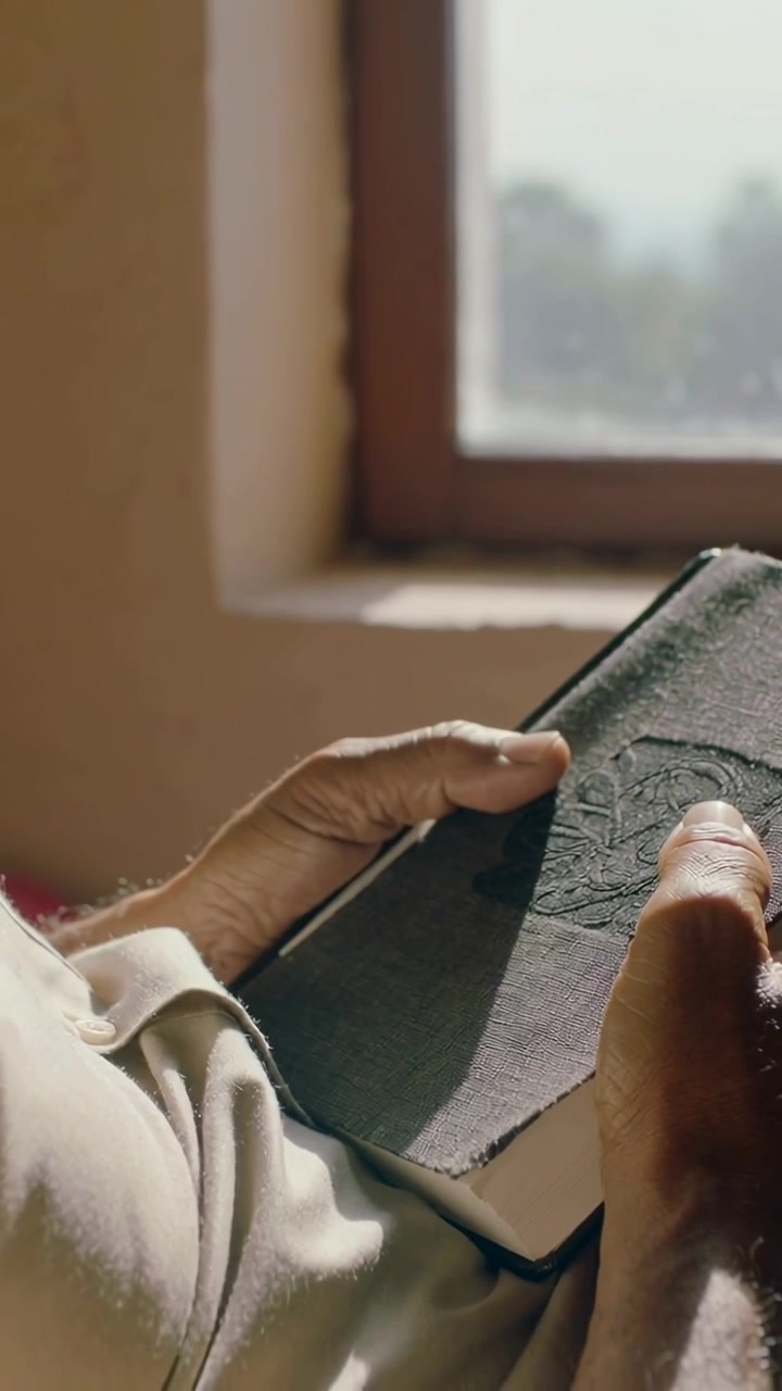 Video verticale: Mani di anziani sistemano un libro di stoffa, toccando la copertina in rilievo vicino a una finestra con polsino beige