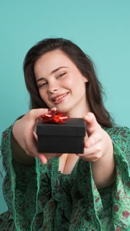 Happy woman giving gift box and looking at camera in blue studio