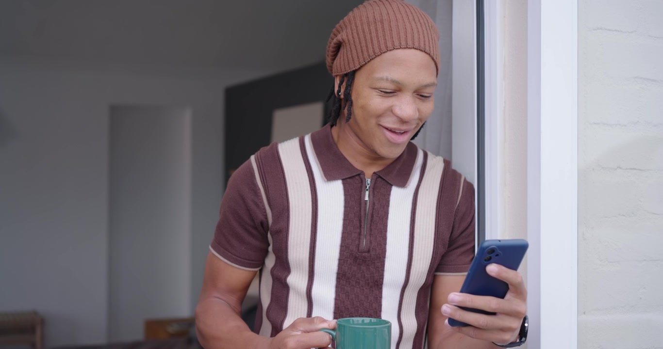 Uomo adulto afroamericano che legge un messaggio sul telefono blu, sorseggiando da una tazza verde e guardando fuori.