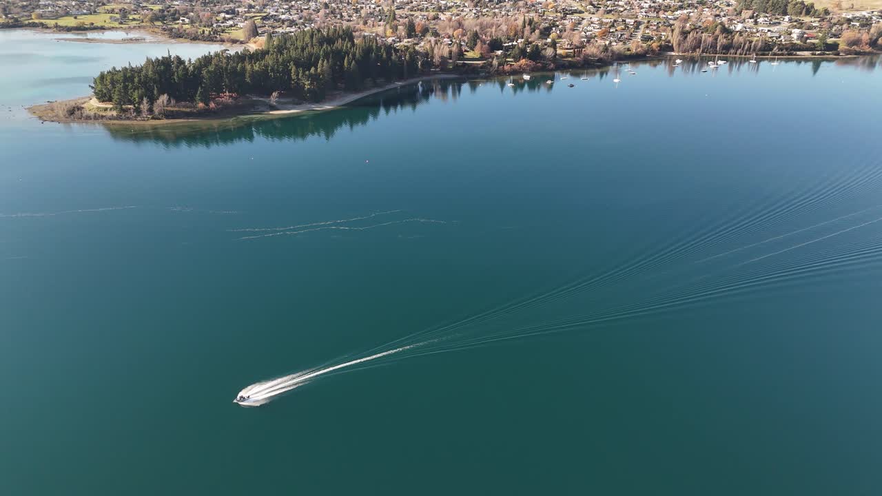 Boat cruising on lake of wanaka during sunny day. Town on island with shoreline. Aerial tracking shot. South Island of New Zealand in summer