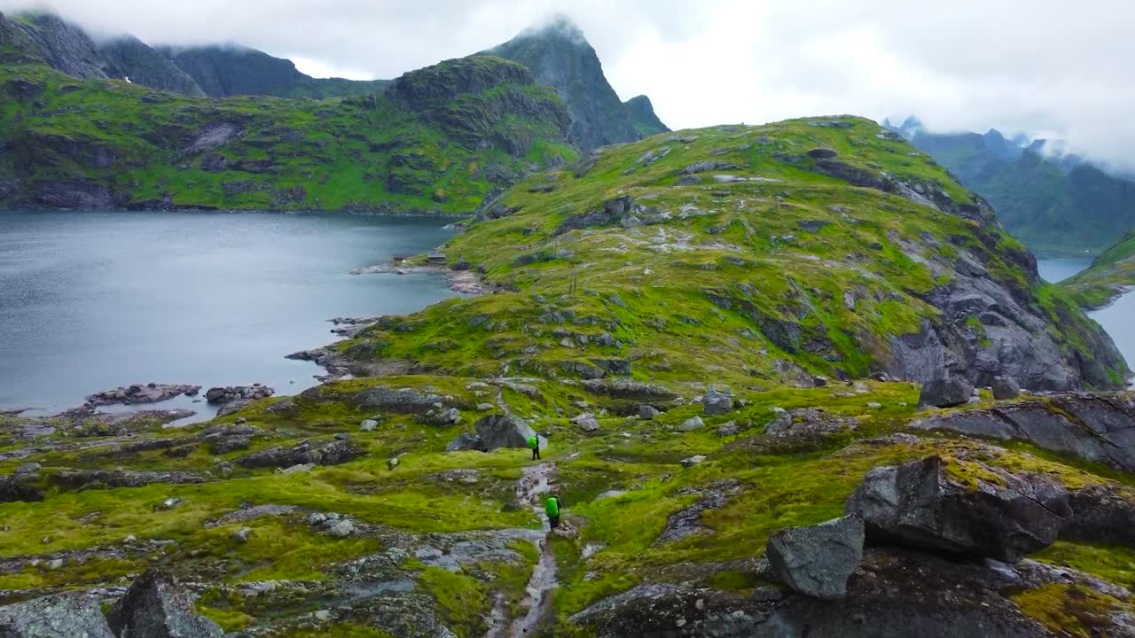 Aerial drone footage flying over a hiking trail with two hikers walking in Lofoten rocky green moss covered mountain terrain in Norway with lakes on both sides during a cloudy day. Mountains visible.