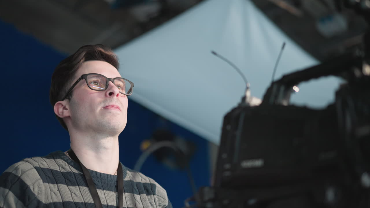 Man standing behind professional video camera in studio setting. Focused crew member operating equipment. Cinematographer adjusting camera during filming session. Filming in production studio