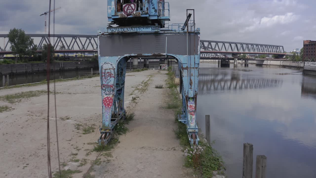 Drone view passing under weathered industrial crane with rusty metal structure and graffiti towards modern bridge crossing river in cloudy day in city