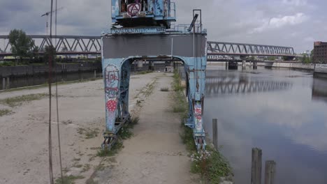drone view passing under weathered industrial crane with rusty metal structure and graffiti towards modern bridge crossing river in cloudy day in city