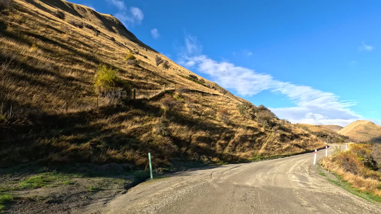 Vehicle travels winding gravel road through sunlit hills, clear sky, and rugged South Island scenery