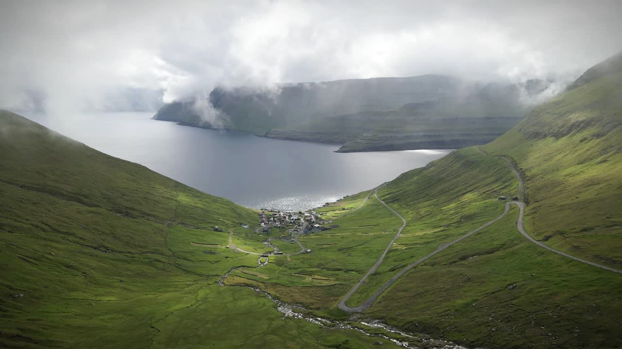 극적인 풍경의 공중 느린 동작, 가파른 초록색 경사 사이에 자리 잡은 매력적인 마을, 조용한 피오르드로 내려가는 휘어지는 도로, 구름이 가득한 하늘 아래, 페로 섬.