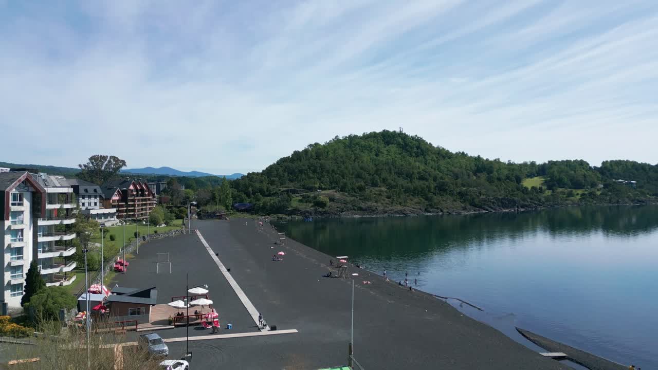 Ascending drone shot over a calm lakeside beach with dark volcanic sand, showcasing waterfront buildings, umbrellas, and a lush green hill across the water in southern Chile