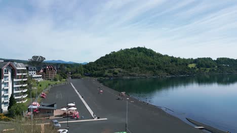 ascending drone shot over a calm lakeside beach with dark volcanic sand, showcasing waterfront buildings, umbrellas, and a lush green hill across the water in southern chile