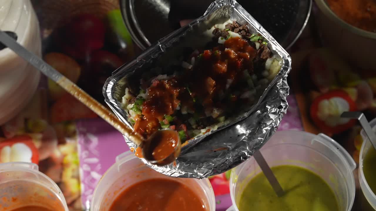 Nærbilledeoptagelse af "papa loca" holdt af en sælger, mens rød sovs generøst øses over fyldet. Den rige sovsfarve og belægning fremhæver intensitet, smagskontrast og præsentationen af street food.