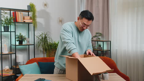 happy chinese man shopper unpacking cardboard box delivery parcel online shopping purchase at home