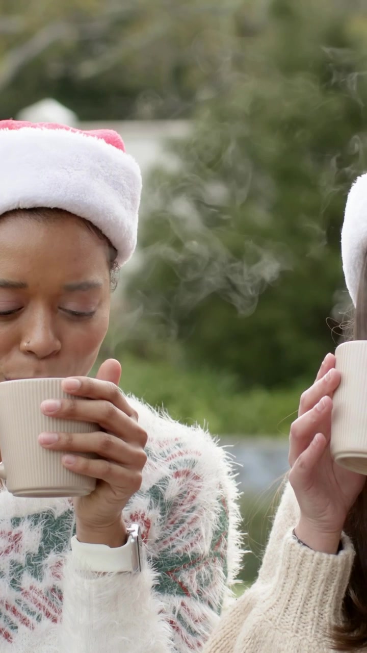 Vidéo verticale : Des tasses s'entrechoquent, la femme de gauche inhale la vapeur et sirote sa boisson pour se réchauffer les mains dans le jardin, portant des bonnets de Père Noël.