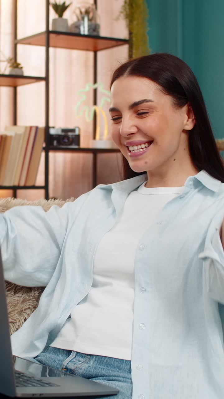 Young woman on sofa raising hands up joyfully celebrating successful finished project on laptop