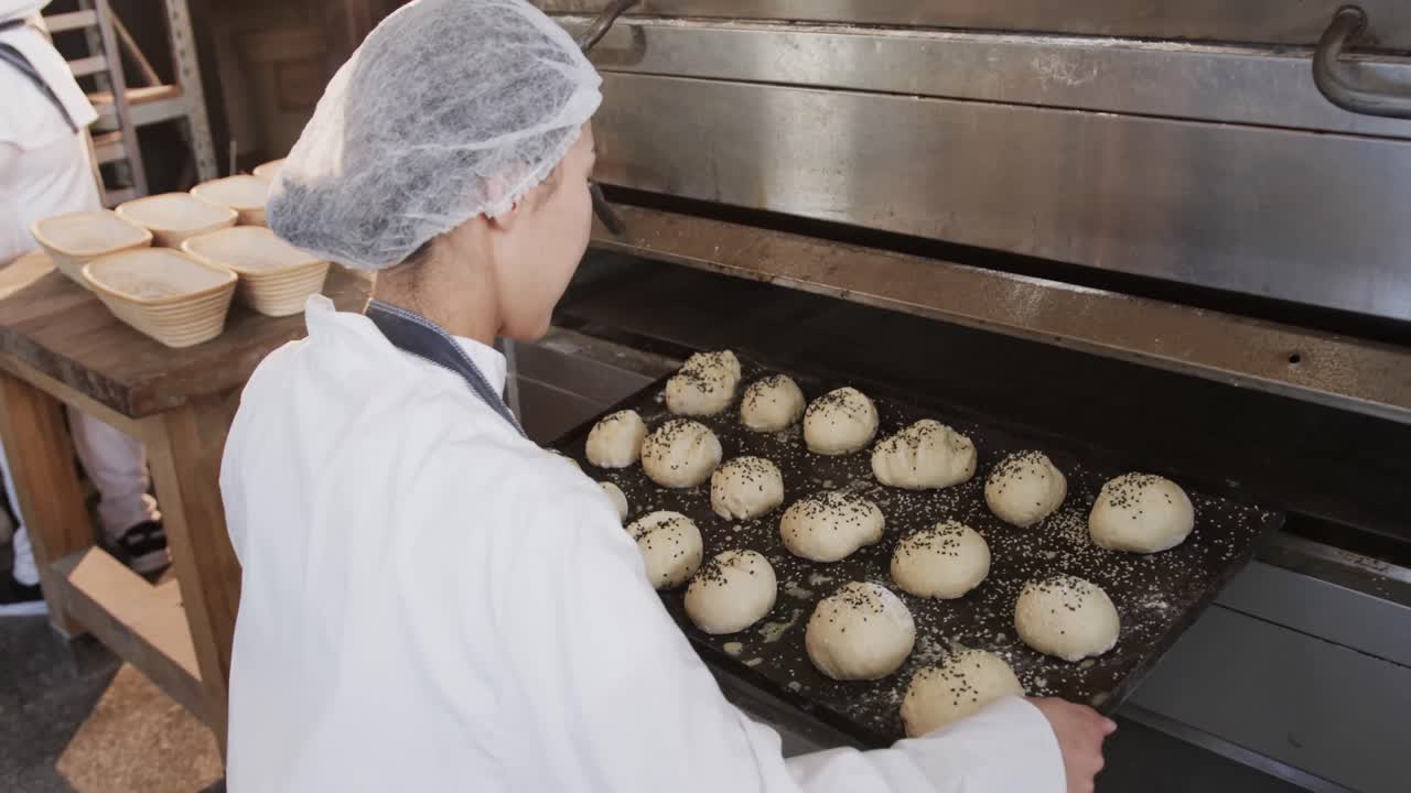 Asian female baker working in bakery kitchen, putting rolls on trays in oven, slow motion