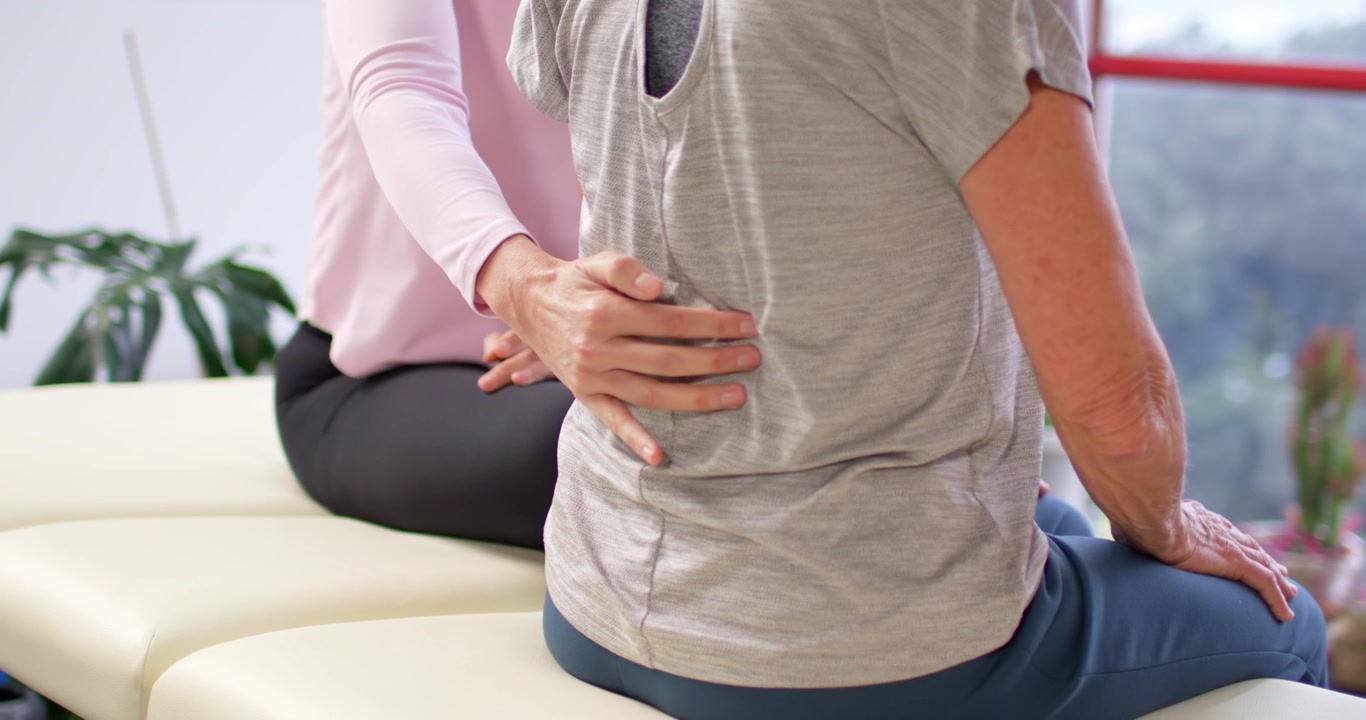 Female clinician palpating senior female patient's lumbar on padded bench assessing alignment