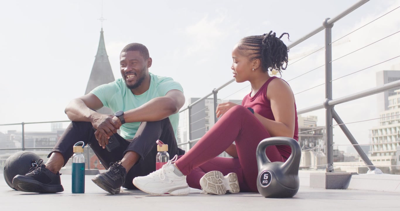African friends sitting rooftop post-workout, woman tying shoe, placing pink bottle by kettlebell