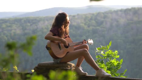 a lady wearing shorts and sneakers seated while playing guitar with forest hills at background. - selective focus