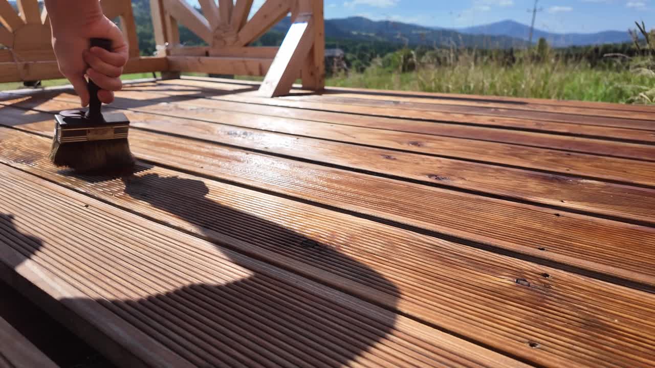 Working man oils varnishing impregnating wooden terrace boards. Decorative railing, serene sky, clouds, and mountain horizon evoke home improvement and outdoor maintenance themes - DIY
