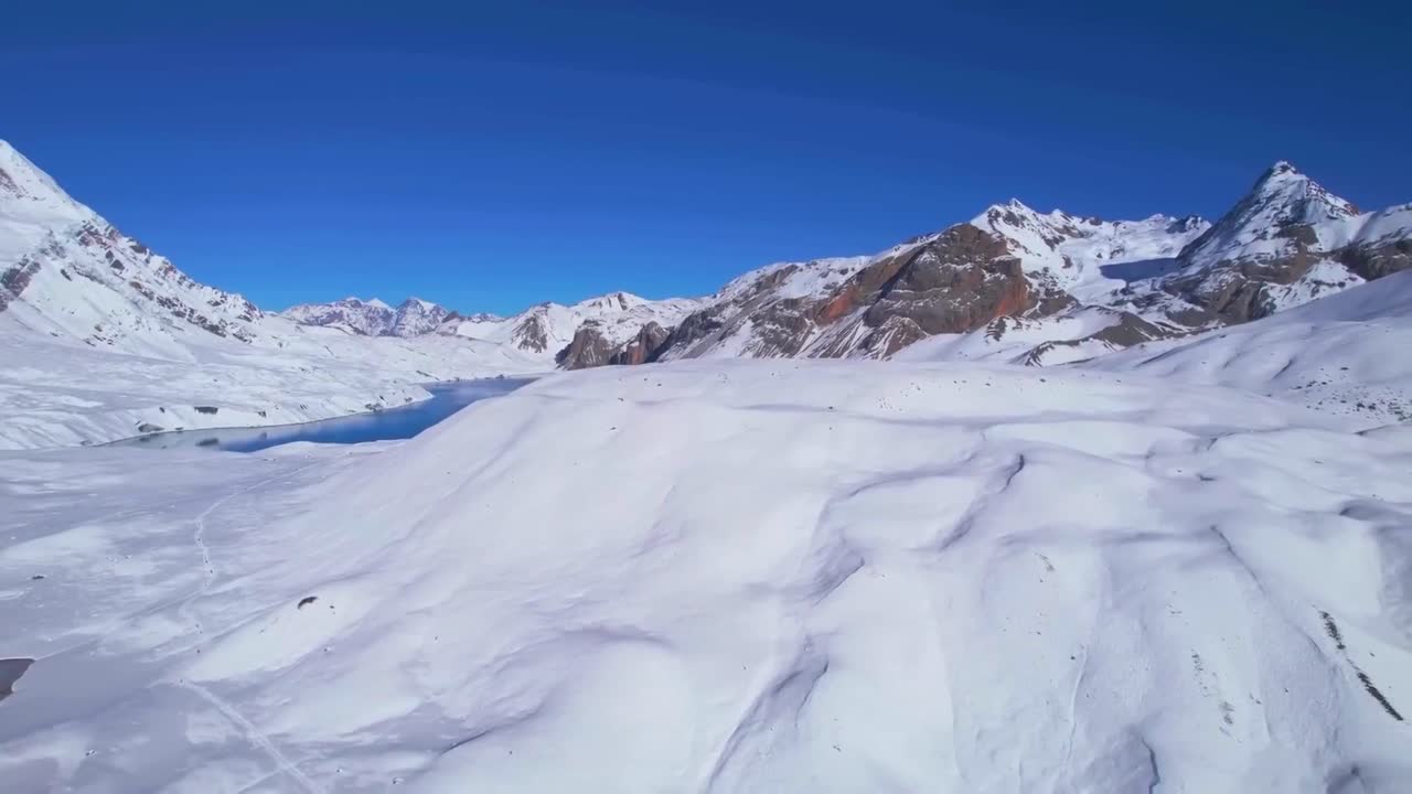 Aerial drone footage slowly flying down while filming Tilicho blue cold icy lake in Nepal during a sunny day. The lake is surrounded by white snow landscape and large tall mountains that have snow on.
