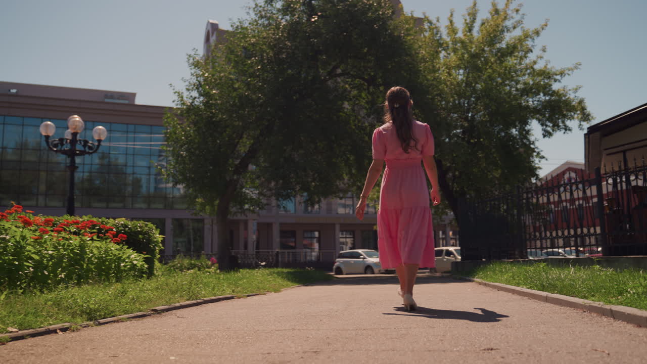 Donna in abito rosa che passeggia lungo un sentiero costeggiato da aiuole fiorite verso un punto di riferimento con cupola di vetro, vista posteriore attraverso un giardino urbano, andatura rilassata ed esplorativa con una leggera brezza, movimento editoriale spontaneo, casual