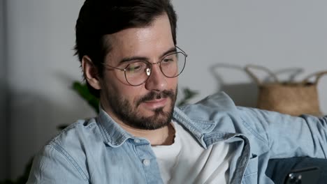 retrato en primer plano de un joven con gafas mirando un teléfono inteligente, aplicaciones en línea, redes sociales, videos y fotos, tendido en un sofá en una habitación en casa